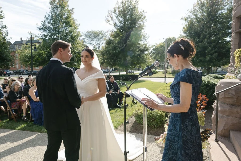 A wedding ceremony outdoors with a bride and groom holding hands and facing each other, while a woman reads from a book at a microphone. Guests are seated in the background on a sunny day.