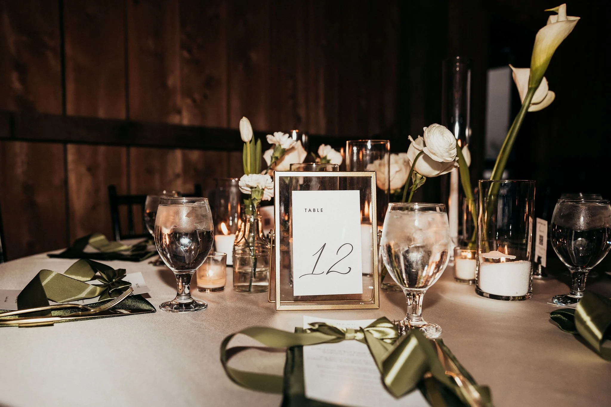 Elegant dining table with white flowers in glass vases, water glasses, candles, a table number card, and green napkin and cutlery, set against a wooden wall background.