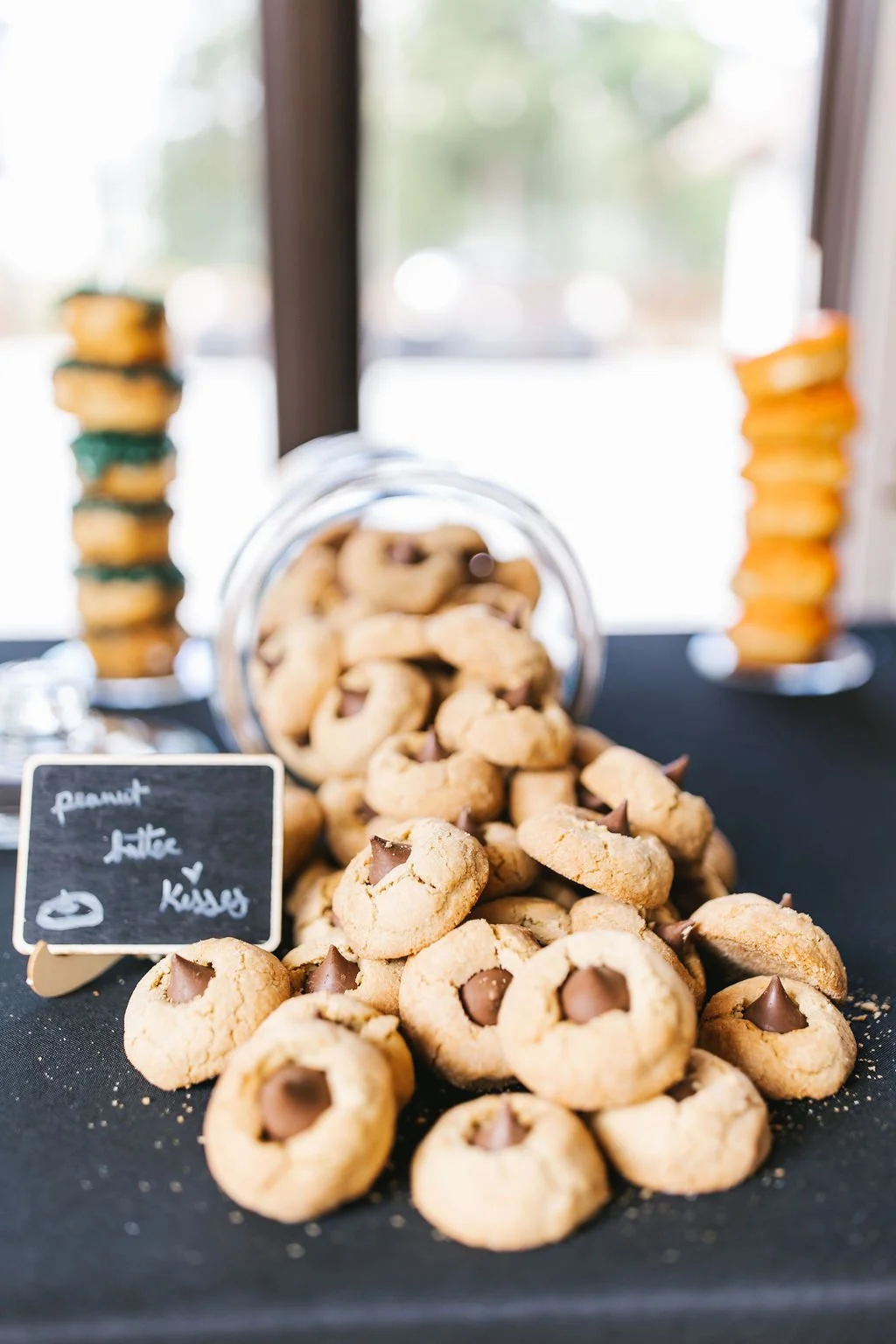 Pile of peanut butter cookies with chocolate chips on black table, with a small blackboard sign reading 'Peanut Butter Kisses' in front of the cookies.
