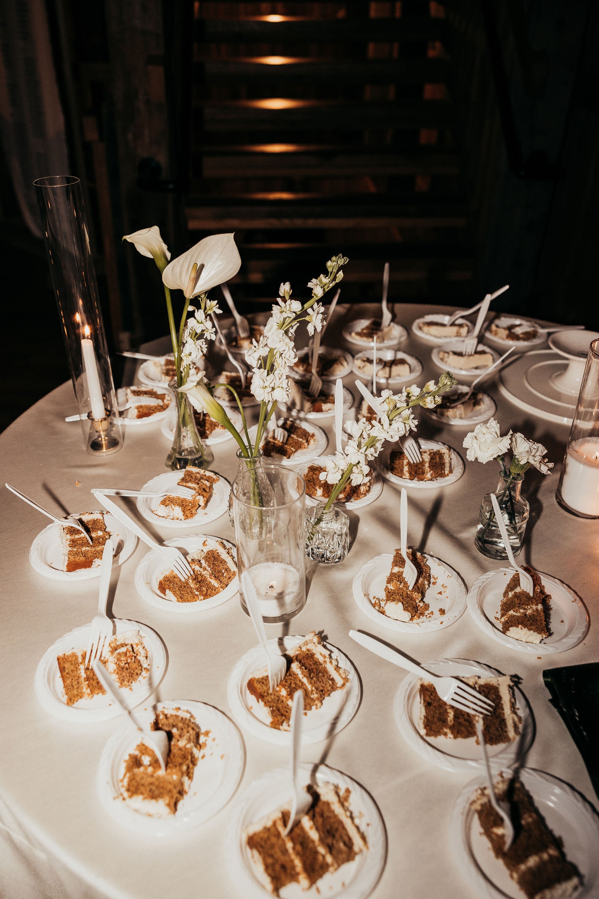 Table with multiple slices of chocolate cake in small paper plates, forks, and decorative flowers in vases, set for a celebration or event.