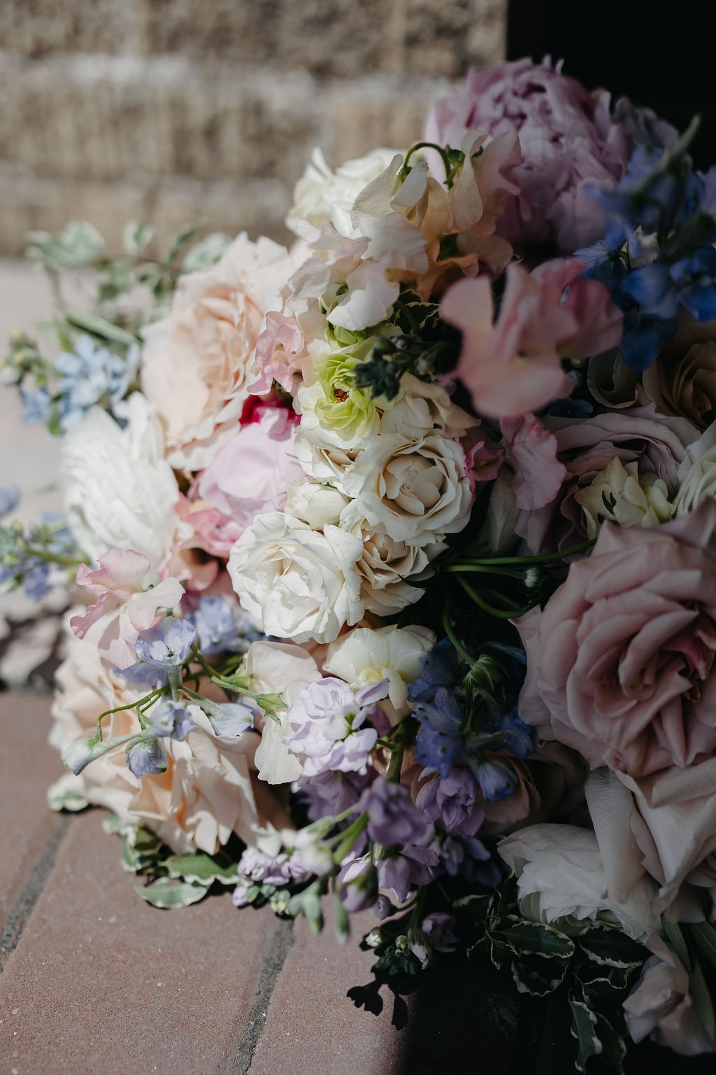 A close-up of a colorful bouquet of flowers with roses, peonies, and other blooms in white, pink, purple, and blue, placed on a brick surface.