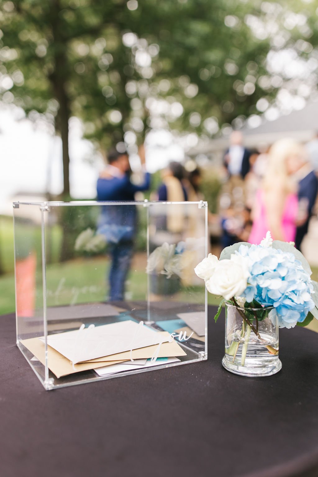 A clear donation box with envelopes inside, placed on a black table next to a vase of blue and white flowers. Blurred group of people gathered outdoors in the background.