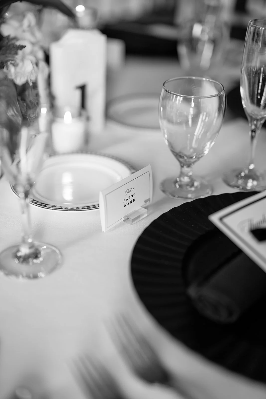 A close-up of a formal dining table with empty wine glasses, a place card reading 'Patti Ward', plates, and a black napkin, arranged for a special event or wedding.