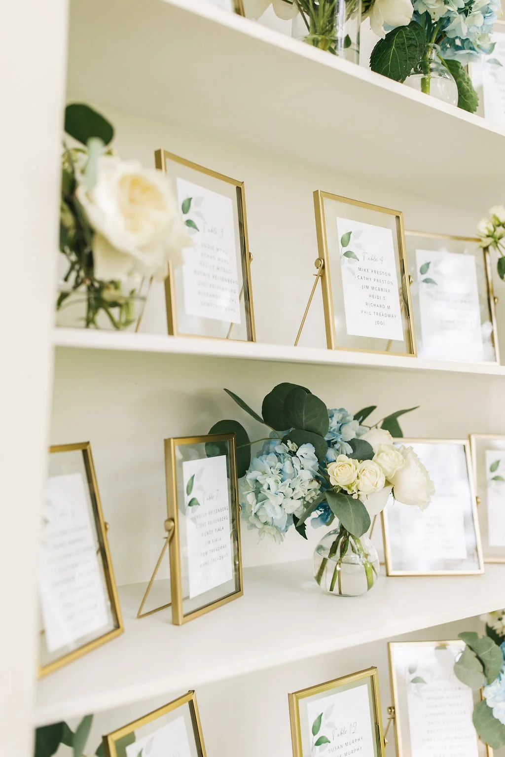 Decorative shelves with picture frames, floral arrangements, and notes for a wedding or event.