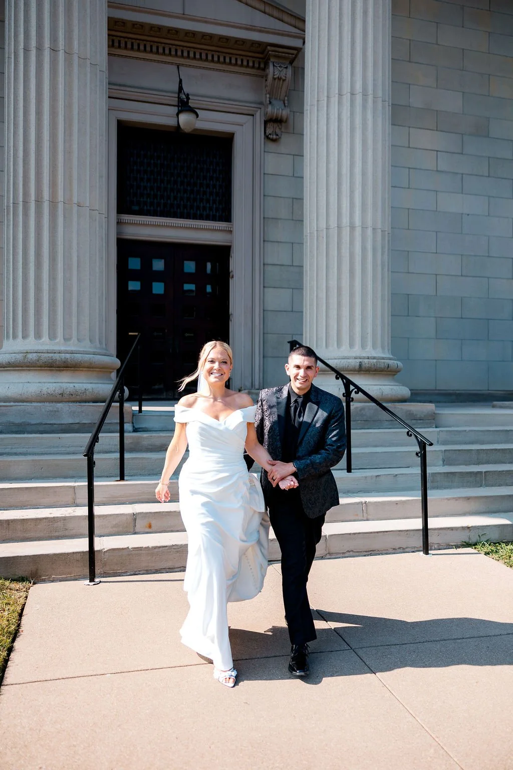 A bride and groom holding hands and smiling as they walk down steps outside a large, classical-style building with columns.