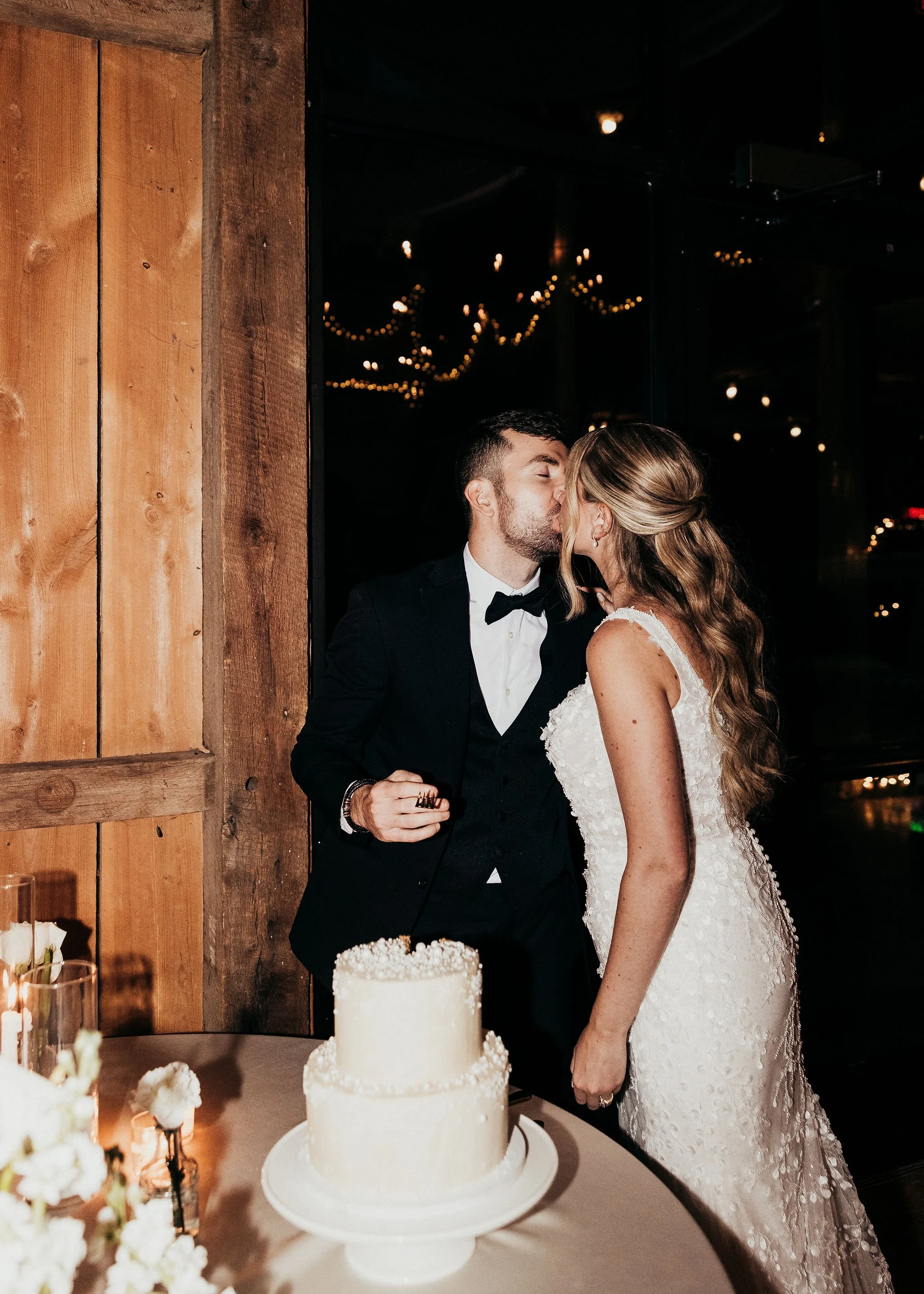 A bride and groom share a kiss in a dimly lit indoor wedding reception. The bride wears a white lace gown, and the groom is in a black tuxedo with a bow tie. A wedding cake is in the foreground, and festive string lights are visible in the background