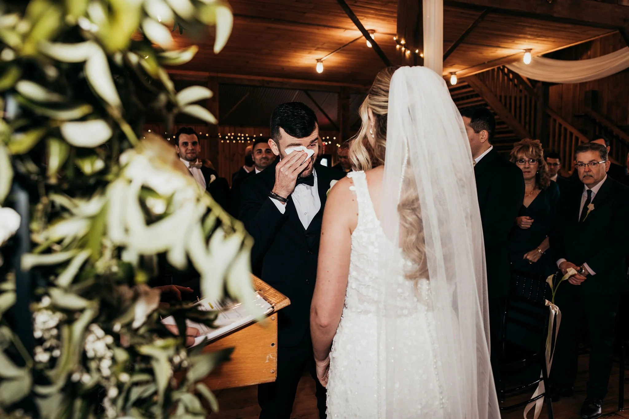 A groom wiping tears at a wedding ceremony in a wooden hall, with guests in the background.