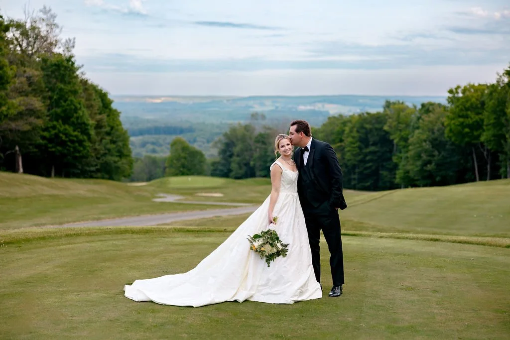 A bride and groom standing on a golf course, with the bride holding a bouquet and the groom leaning in to kiss her on the forehead, surrounded by green trees and a scenic landscape.