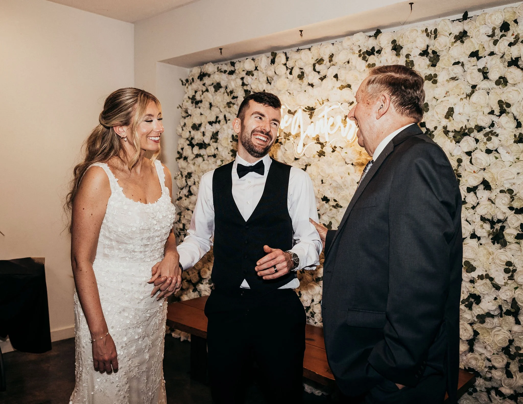 A bride and groom at their wedding ceremony exchanging vows with an officiant in front of a wall of white roses.