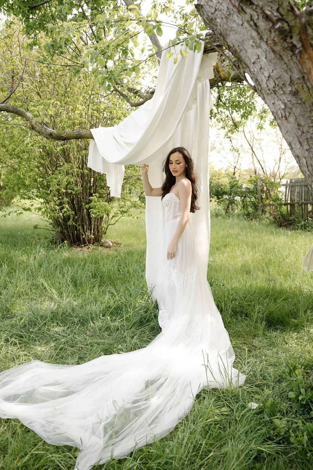 A woman in a white wedding dress standing outdoors in a grassy yard, holding a piece of white fabric that is draped from a tree branch, with sunlight filtering through the leaves.