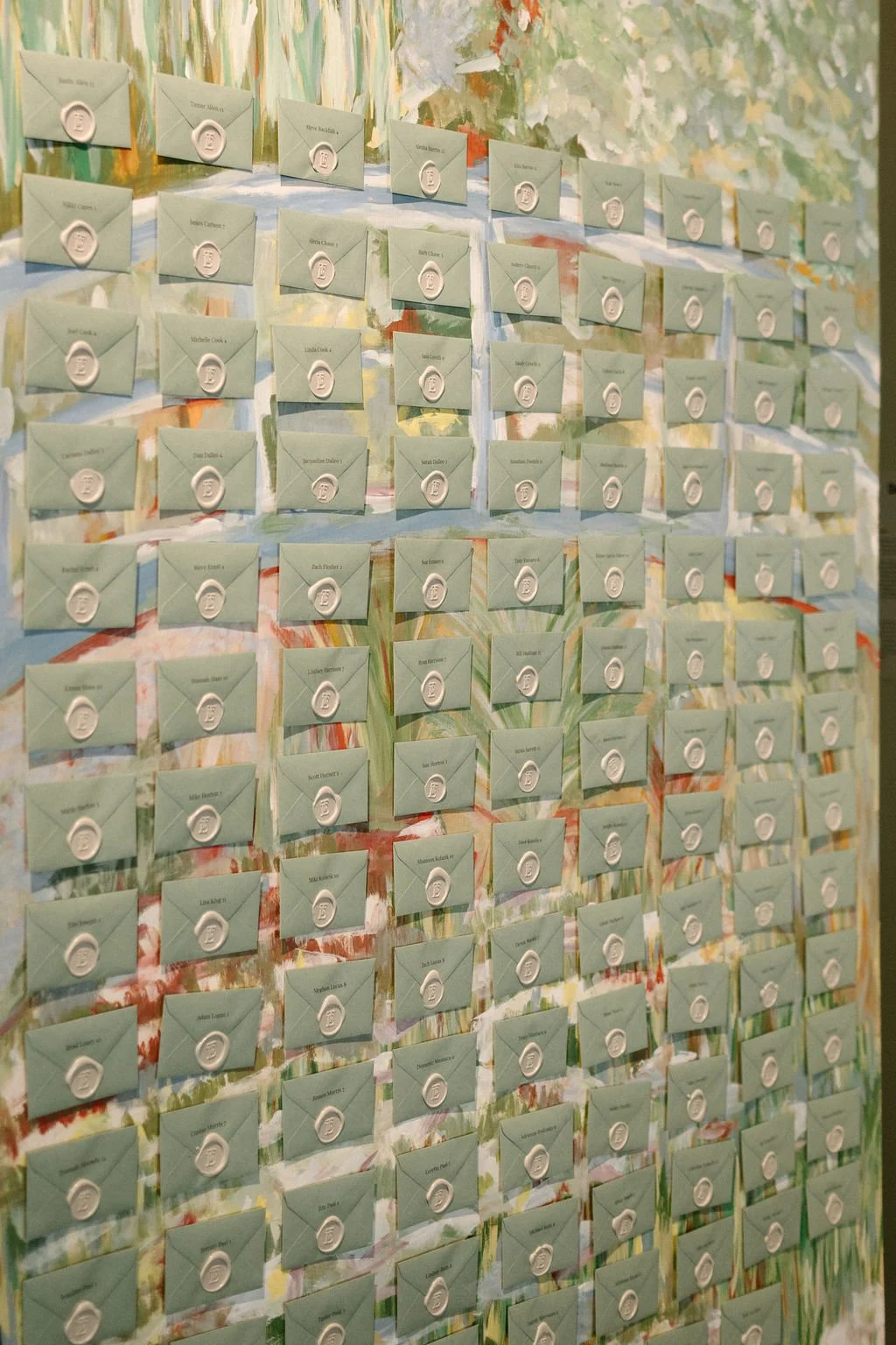 A wall decorated with multiple rows of small, light green envelopes each sealed with a white wax stamp, with names written on the envelopes, against a colorful, abstract, painted background.