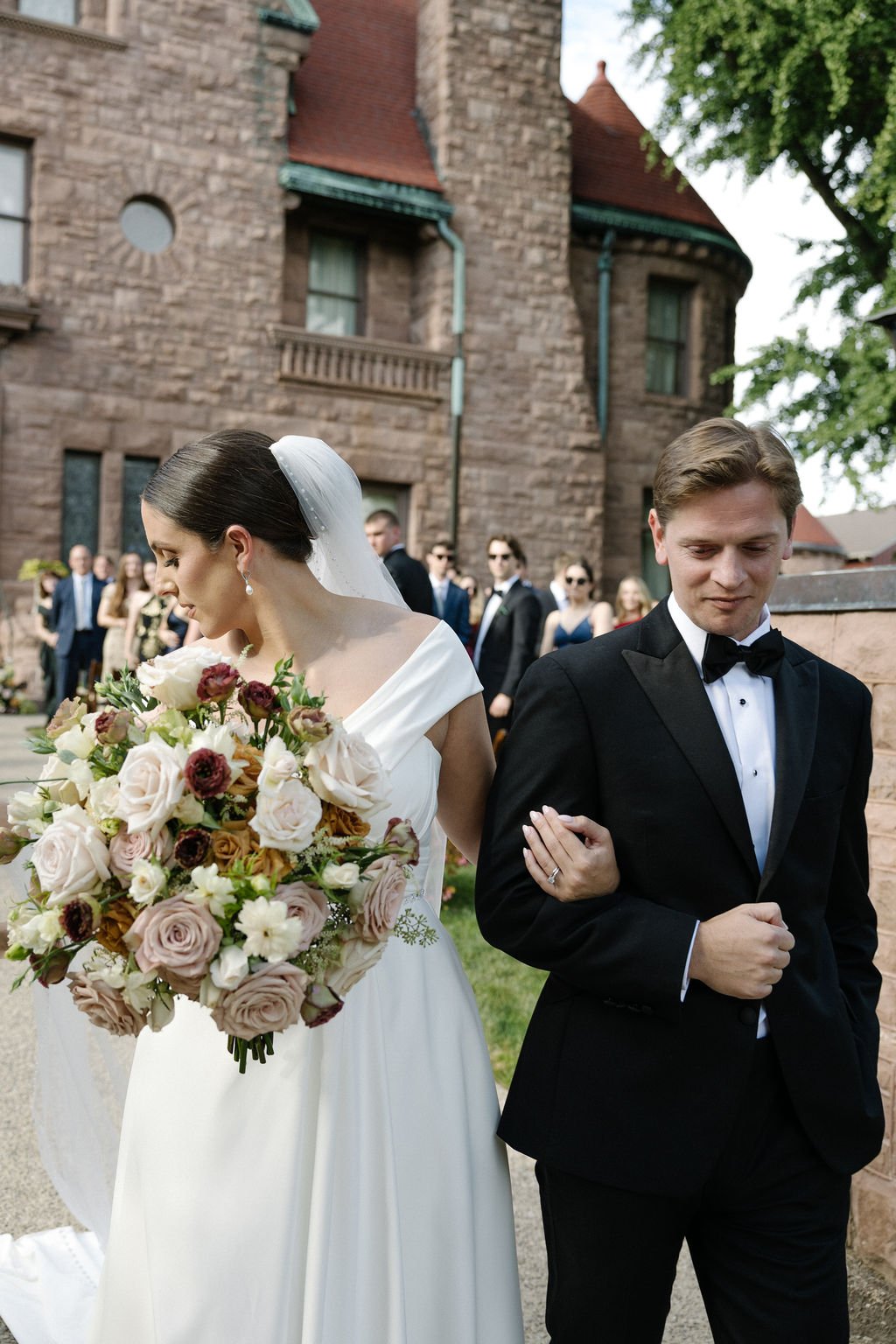 A bride in a white wedding dress holding a bouquet of flowers stands beside a groom in a black tuxedo at an outdoor wedding ceremony. Wedding guests are in the background, and a historic stone building is behind them.