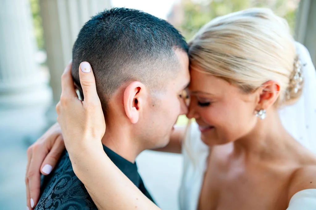 Close-up of a bride and groom with foreheads touching, embracing outdoors during a wedding ceremony, with natural light and columns in the background.