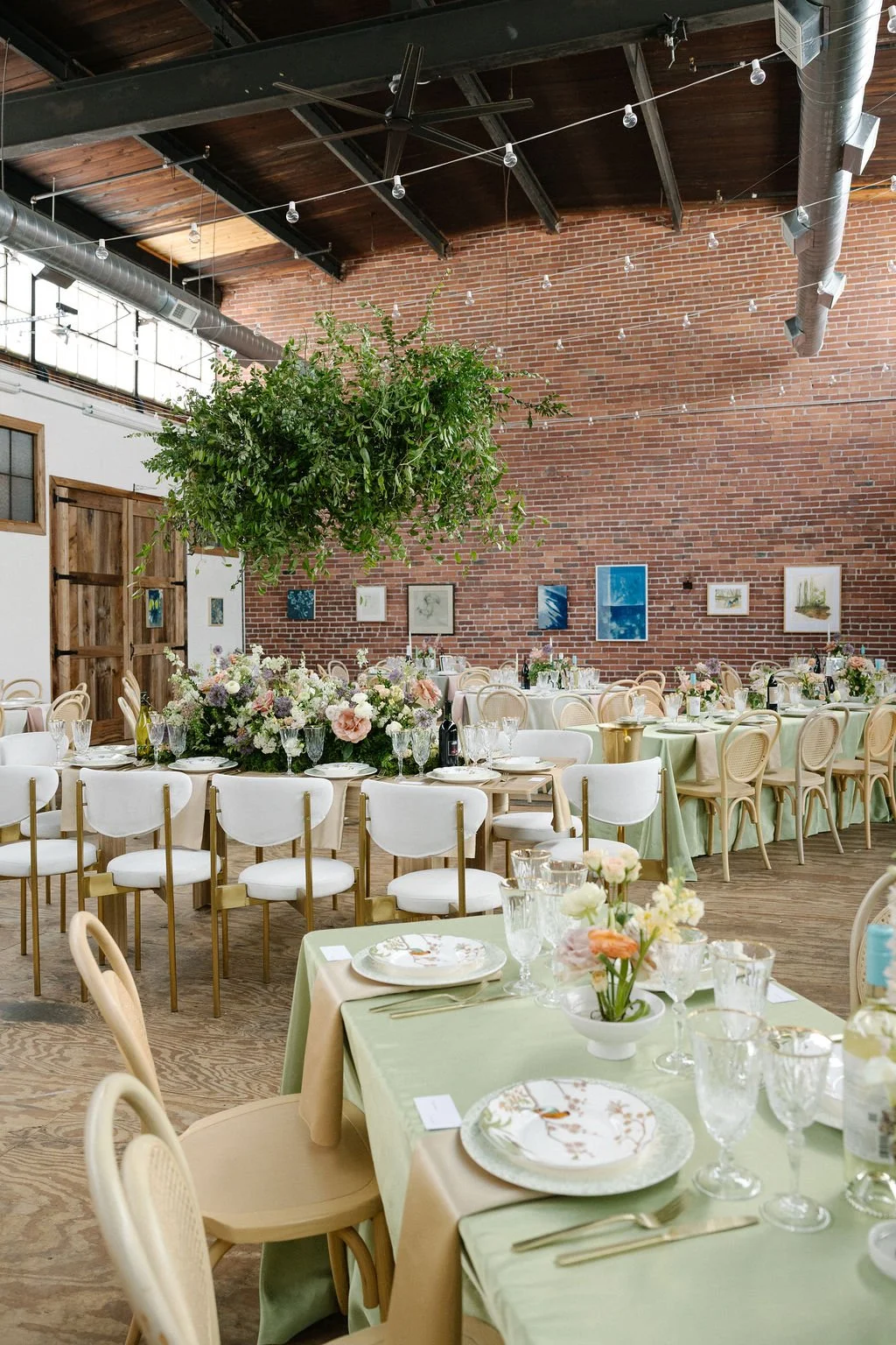 Decorated indoor event space with tables set for a formal meal, floral centerpieces, and a large hanging greenery. Brick wall with art, string lights, and a wooden door are visible.