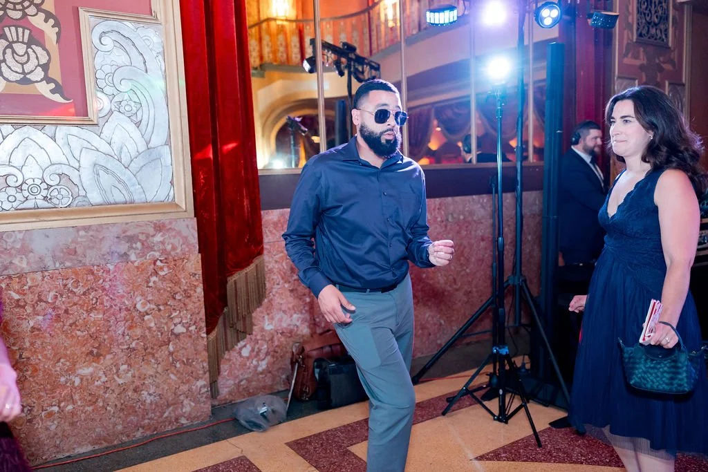 A man with sunglasses and a beard dancing at a formal event with a woman in a dark blue dress holding a clutch, in a decorated indoor venue with colorful lighting.