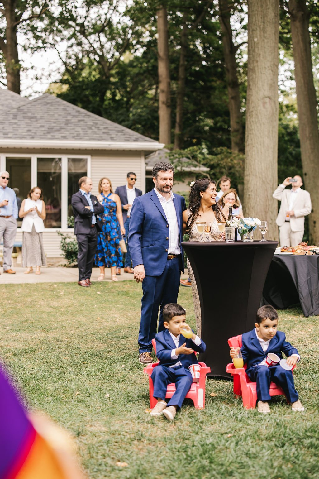 A wedding reception outdoors with guests, including children in suits seated on small chairs, and a couple standing near a table with drinks and flowers, all set in a green yard with trees and a house in the background.