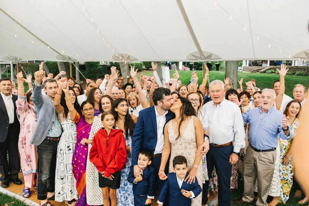 A large group of people celebrating under a tent, some cheering, with a couple kissing in the center, indicating a joyful event like a wedding.