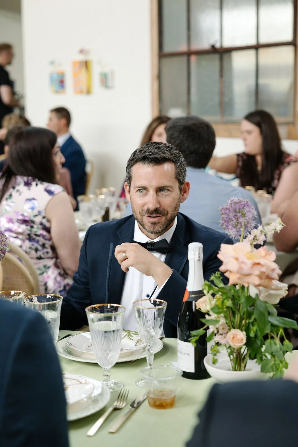 A man in a tuxedo with a bow tie, sitting at a table during a formal event, surrounded by floral arrangements and glassware.