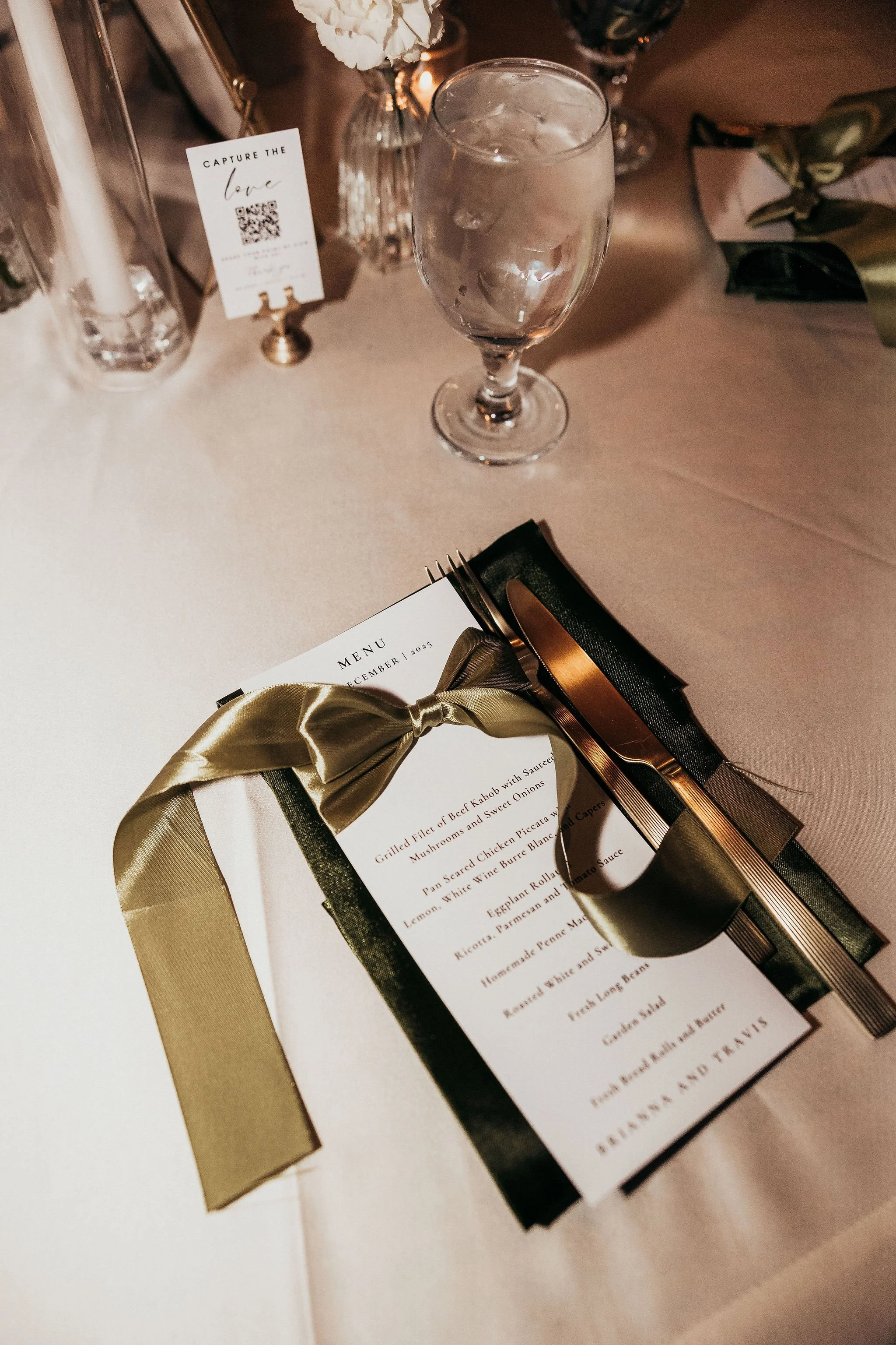 A wedding or special event table setting featuring a glass of water with ice, a menu, and a gold-wrapped gift tied with a gold ribbon, with floral arrangements and candles in the background.