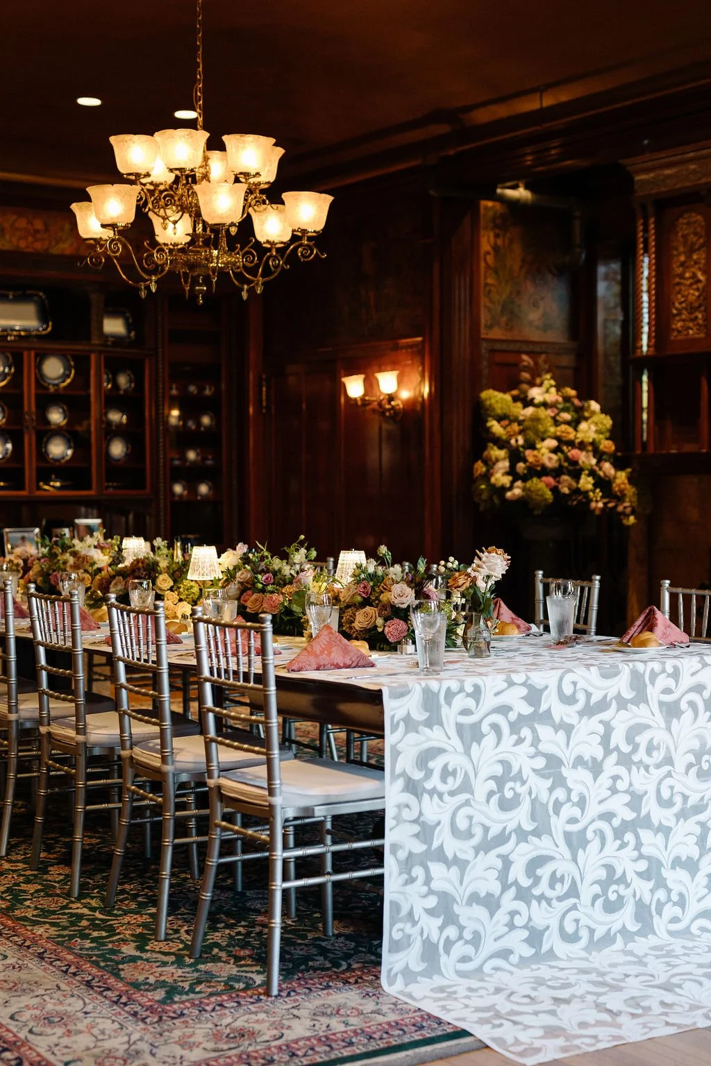 Elegant banquet table setup with floral centerpieces, pink napkins, and glassware in a dimly lit room with chandeliers and wooden wall paneling.