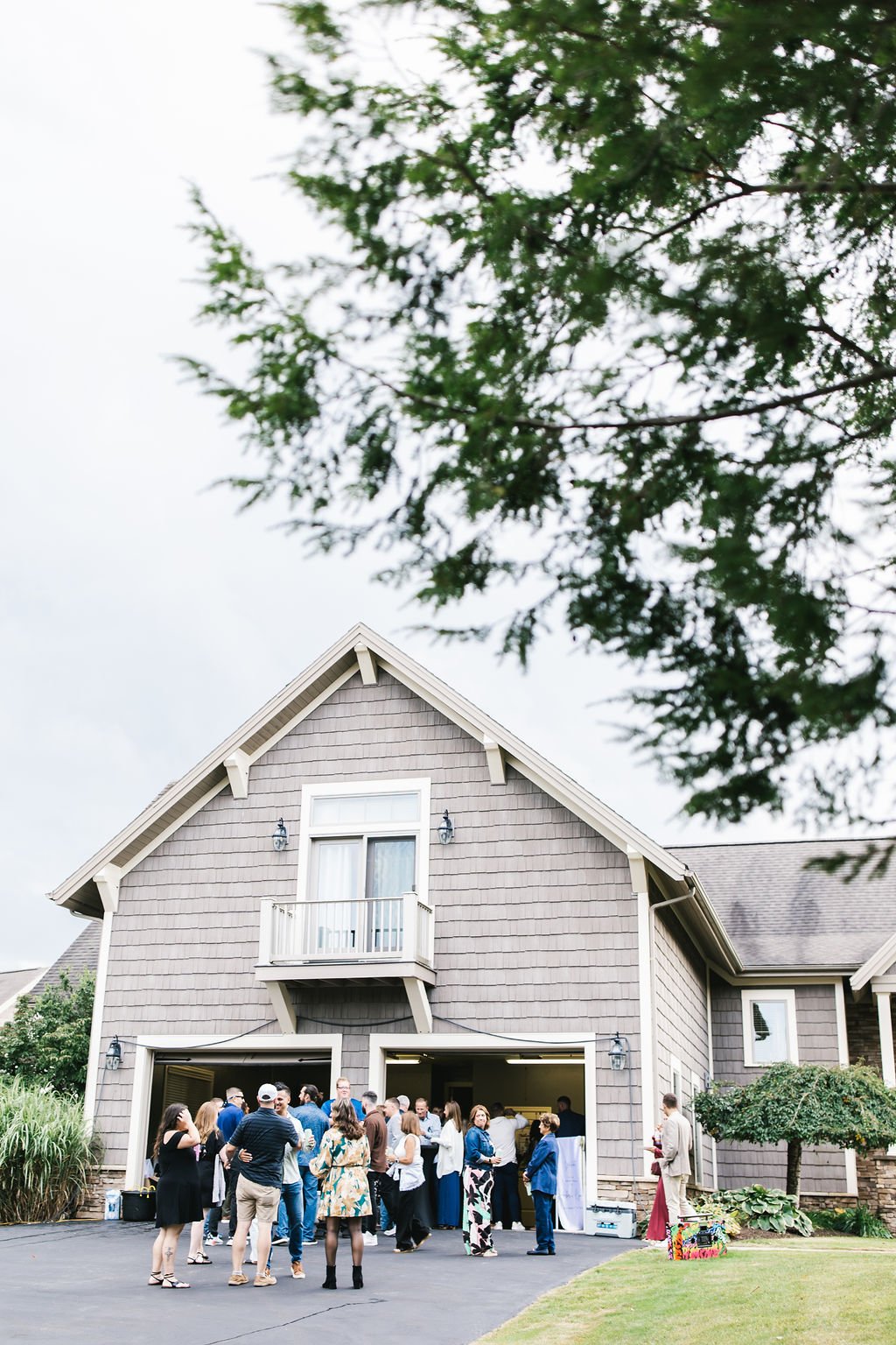 People gathered outside a gray two-story house with an open garage, some standing and chatting, under a cloudy sky, with trees in the background.