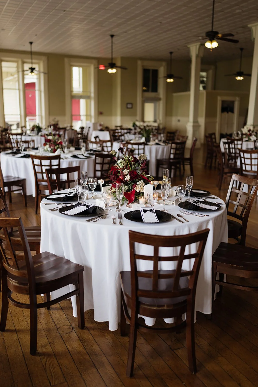 Elegant banquet hall with round tables covered in white tablecloths, each set with black placemats, silverware, wine glasses, candles, and floral centerpieces featuring red, white, and green flowers. Wooden chairs surround the tables. The hall has la