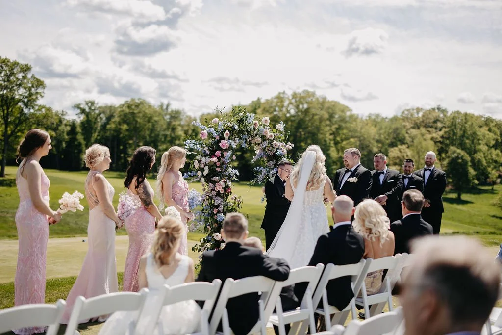 A wedding ceremony outdoors on a sunny day, with a bride and groom exchanging vows under a floral arch, surrounded by bridesmaids in pink dresses and groomsmen in tuxedos, seated guests, and lush green scenery.