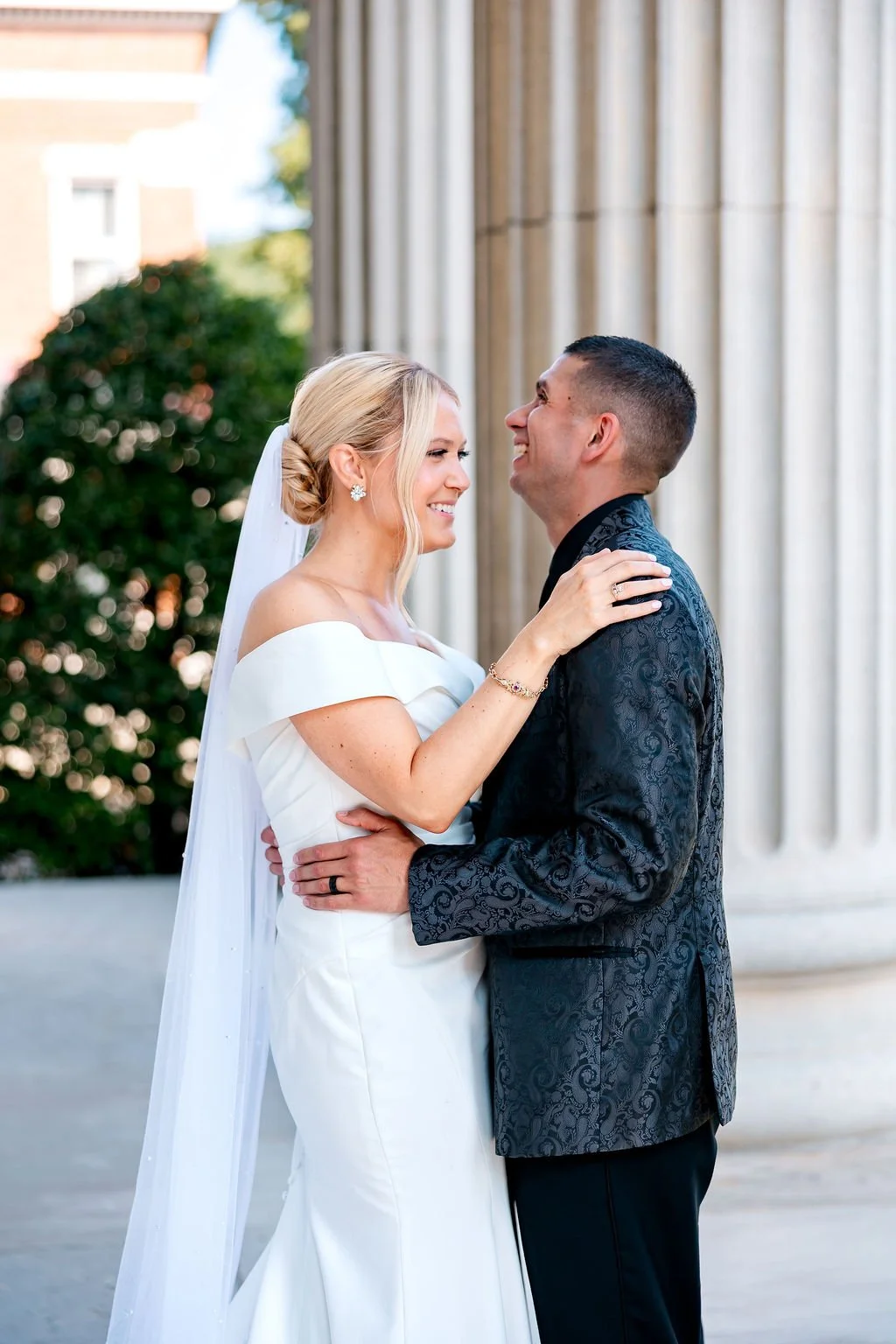 A bride and groom smiling at each other outdoors in front of classical columns during their wedding.