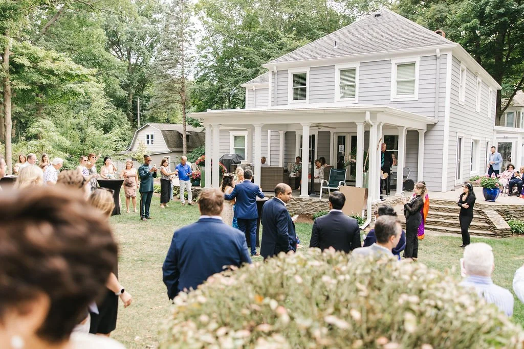 People gathered outdoors at a house party or event, standing and sitting on a lawn in front of a white two-story house with a porch, surrounded by trees.