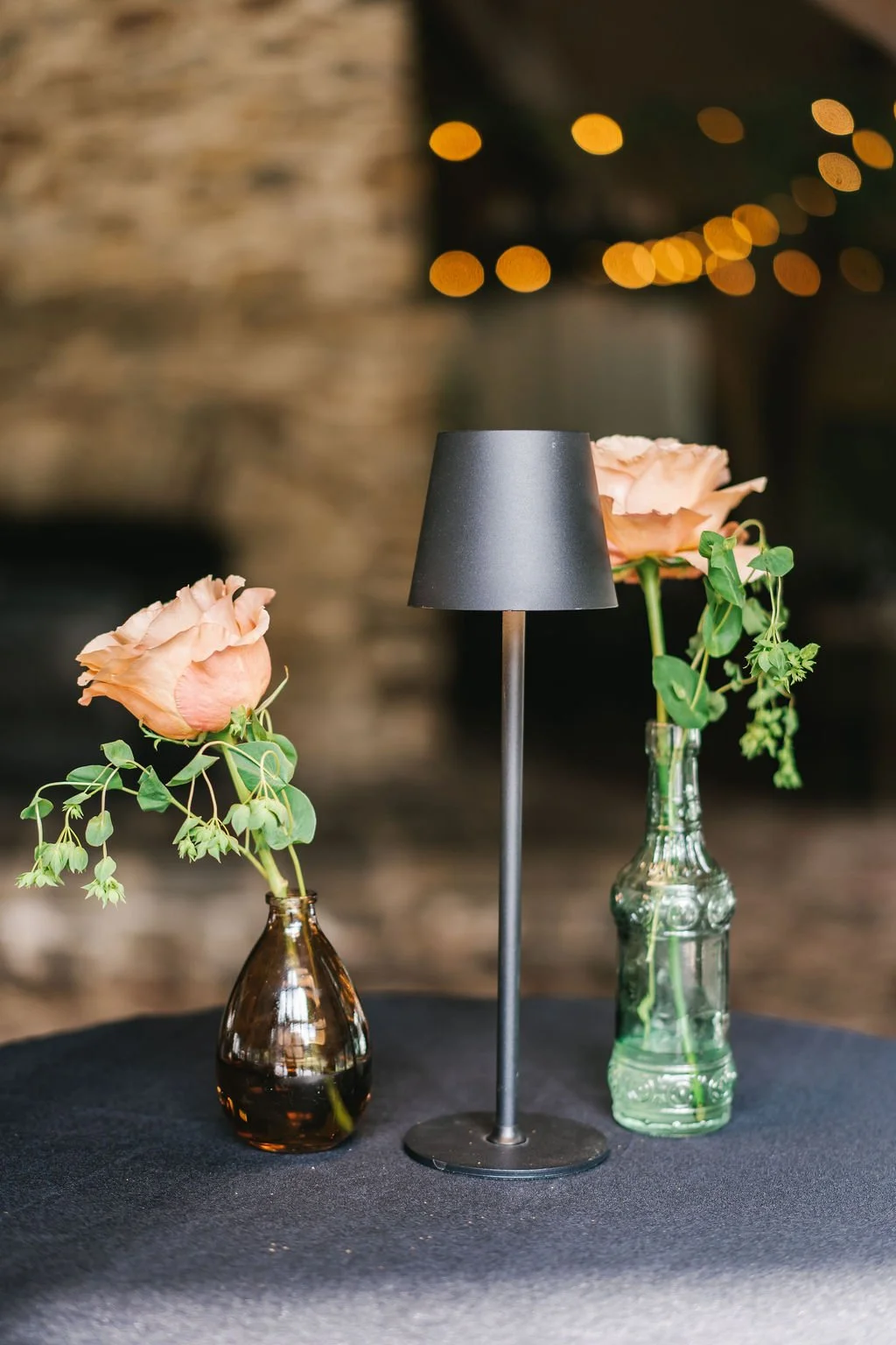 Two vases with flowers, a black table lamp, and a dark table surface with blurry warm lights in the background.
