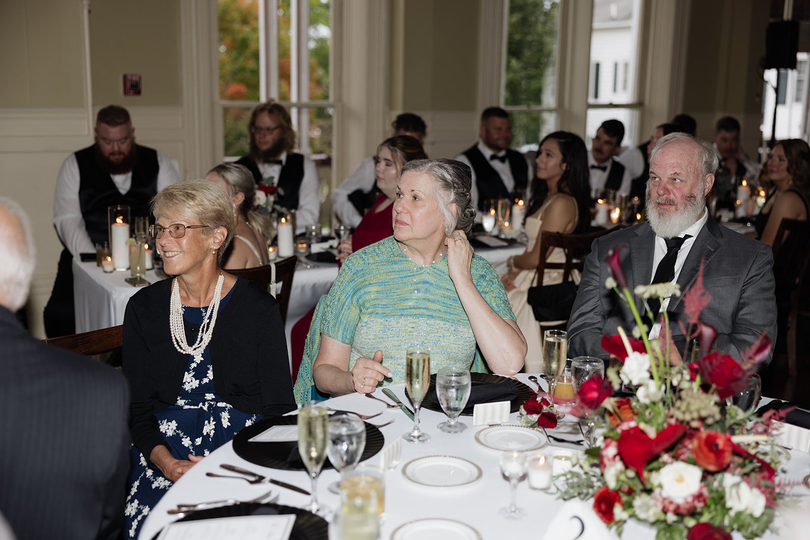 Guests sitting at a decorated banquet table during a formal event or celebration, with candles and floral centerpieces, in a well-lit room with large windows and greenery outside.