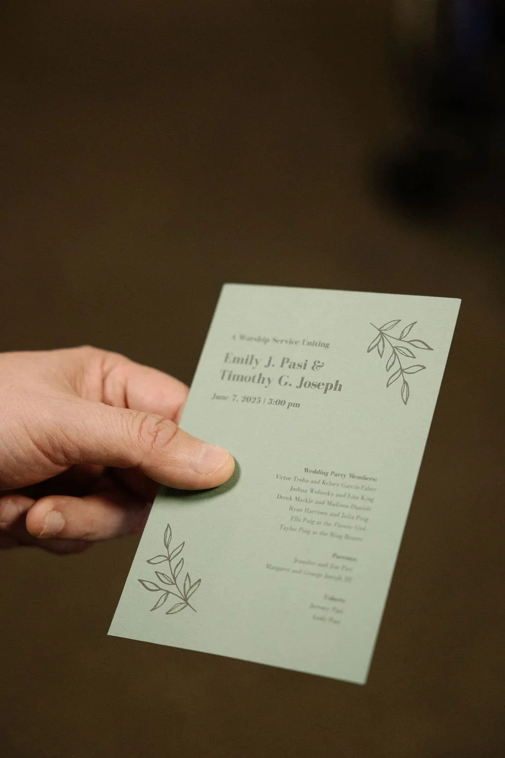A person holding a wedding program with greenish paper, black text, and decorative leaf illustrations on a dark background.