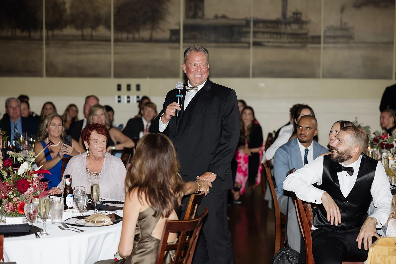 Man in black tuxedo giving a speech at a formal dinner event, surrounded by seated guests in elegant attire, with a large mural in the background.