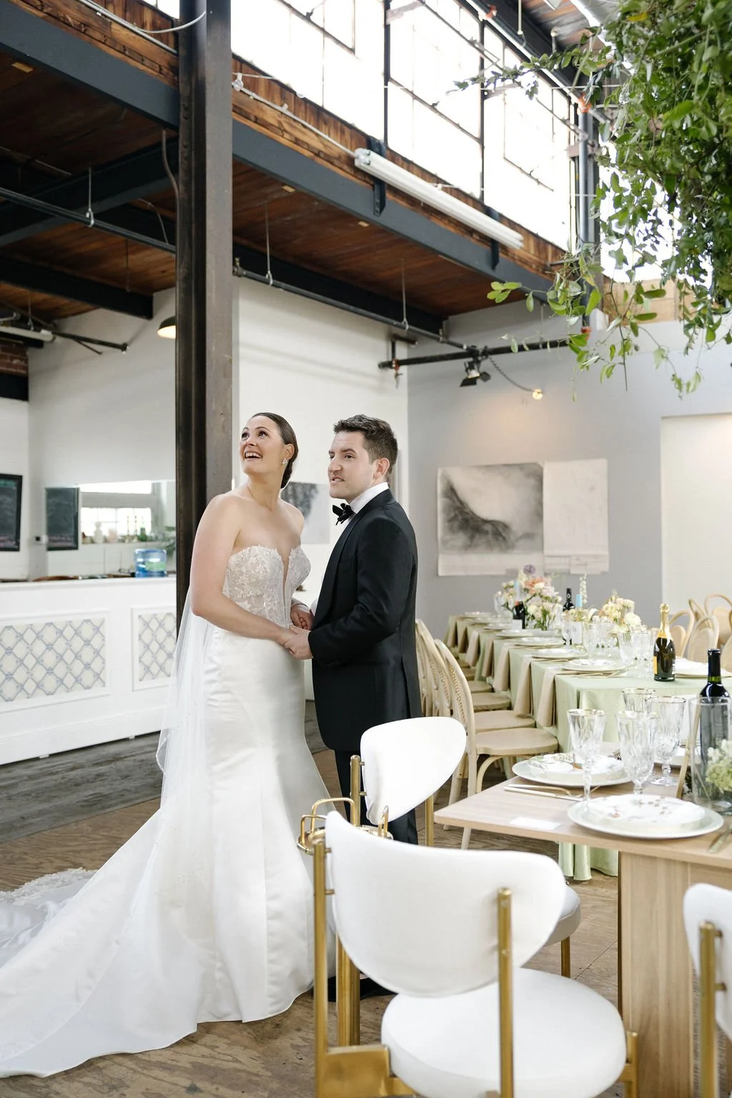 Bride and groom holding hands inside a decorated wedding reception venue, with tables set with glassware and floral arrangements.