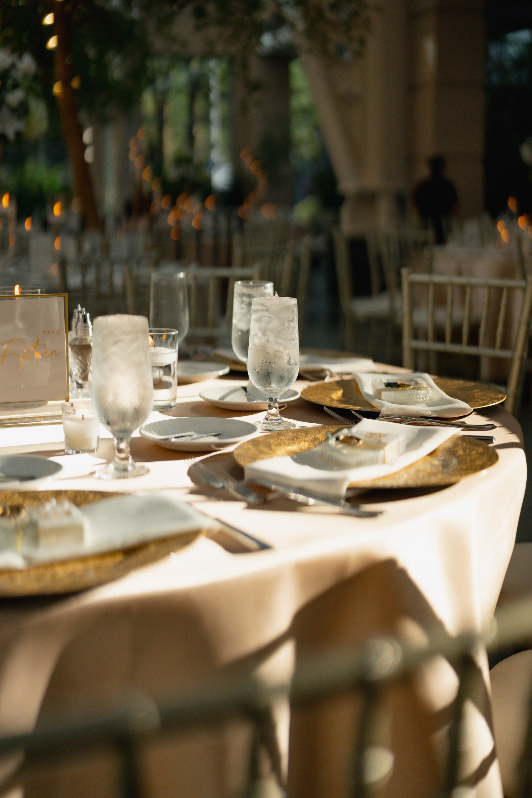 A blurred outdoor dining scene with tables set with glassware, plates, candles, and decorative placemats, with a person standing in the background near a large structure and trees.