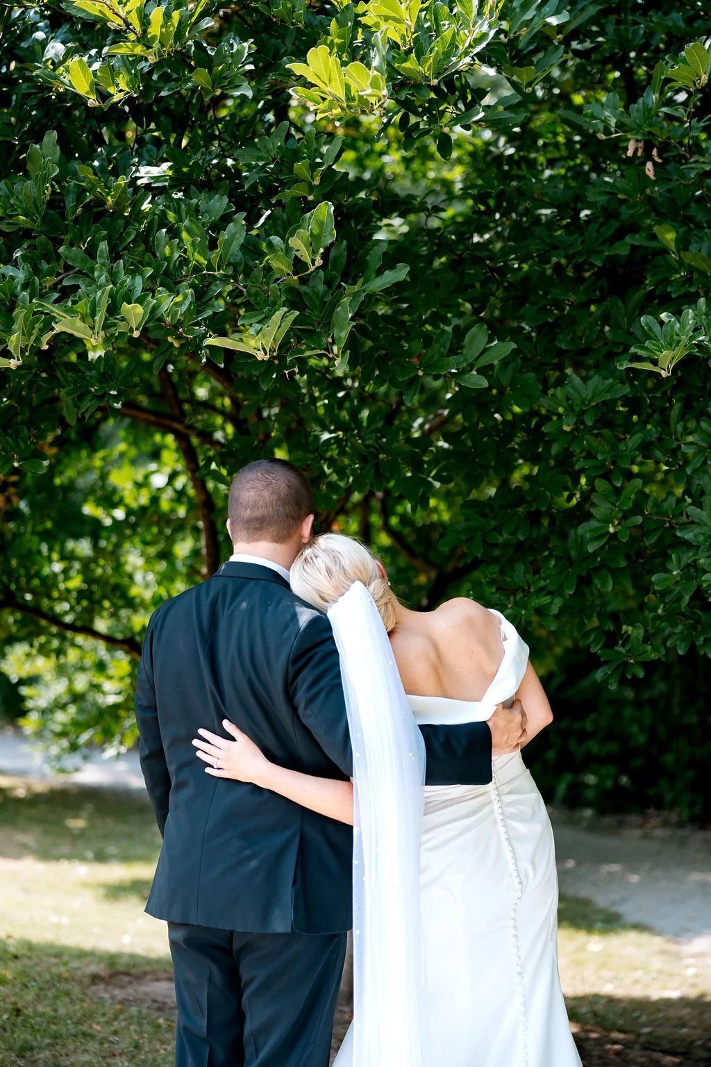A bride and groom embracing outdoors under a lush green tree. The groom is wearing a black suit and the bride is wearing a white wedding dress with a long veil.