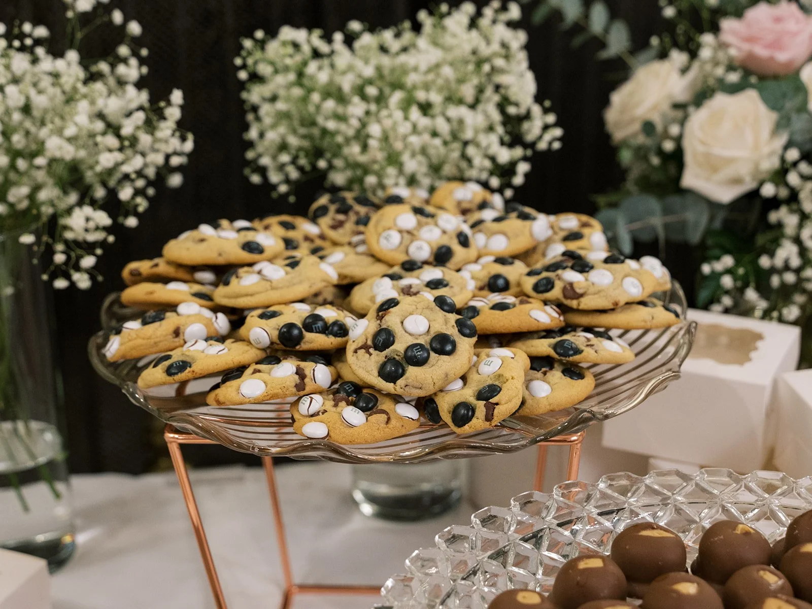 A tray of chocolate chip cookies decorated with white and black candies, with flowers in the background.