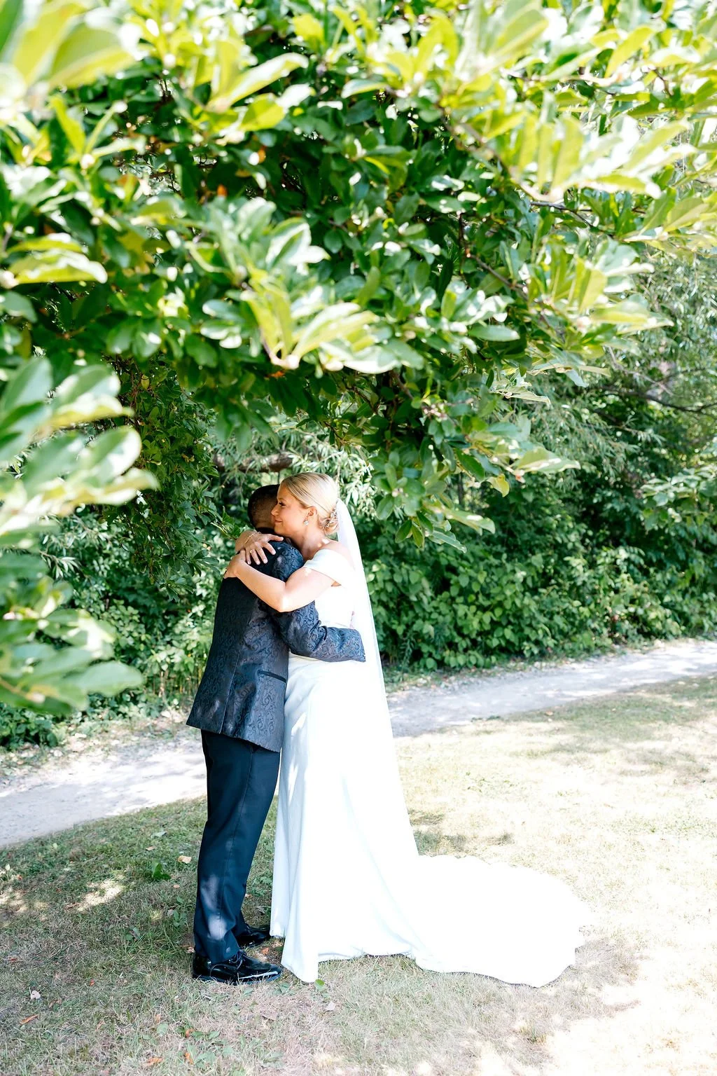 A bride and groom share a kiss outdoors beneath lush green trees.
