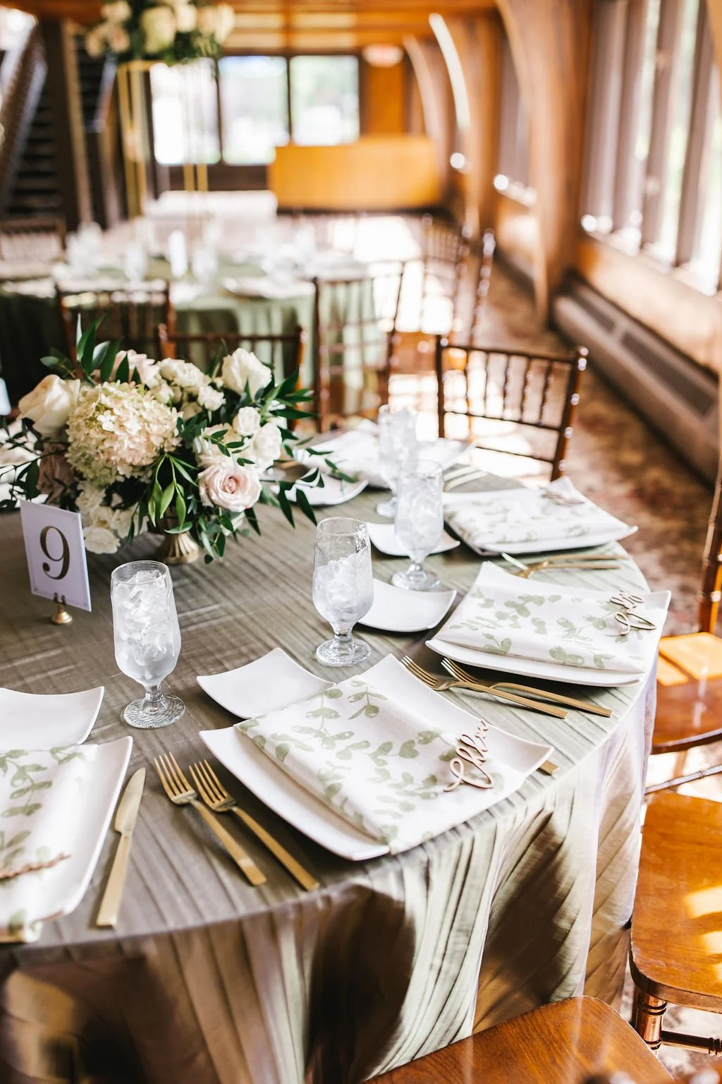 A beautifully arranged wedding or event table decorated with a floral centerpiece, white plates, gold cutlery, patterned napkins, and water glasses, set in an elegant wooden room with natural light streaming through the windows.