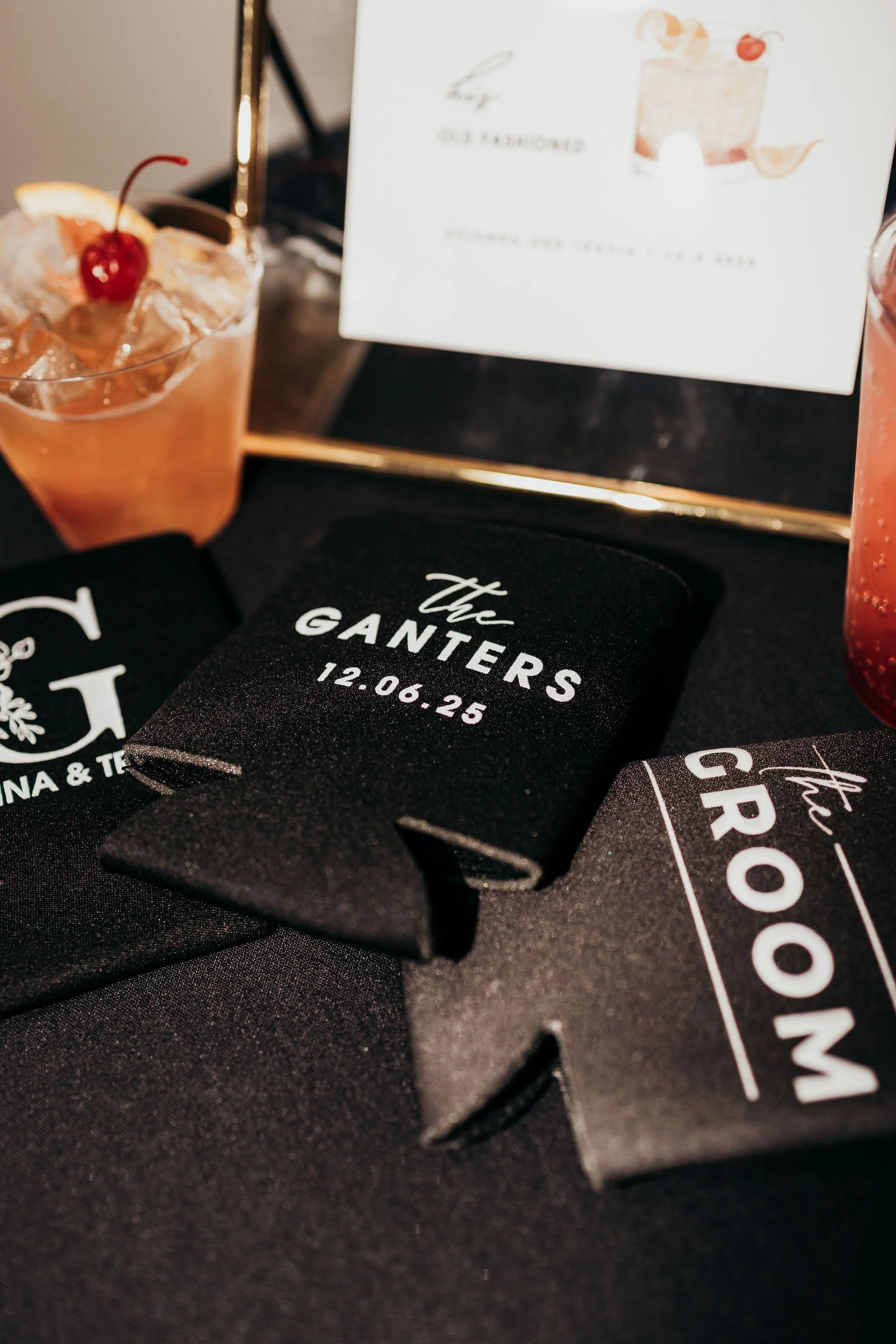 Black fabric items with white text and a printed wedding date, along with promotional material and drinks with cherries and fruit garnishes on a black surface.