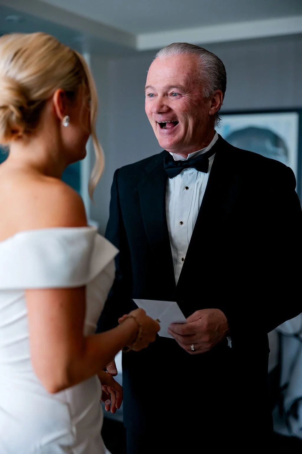 An elderly man in a black tuxedo and bowtie smiling and holding a card, speaking to a woman in a white dress with off-the-shoulder sleeves, during a wedding ceremony.