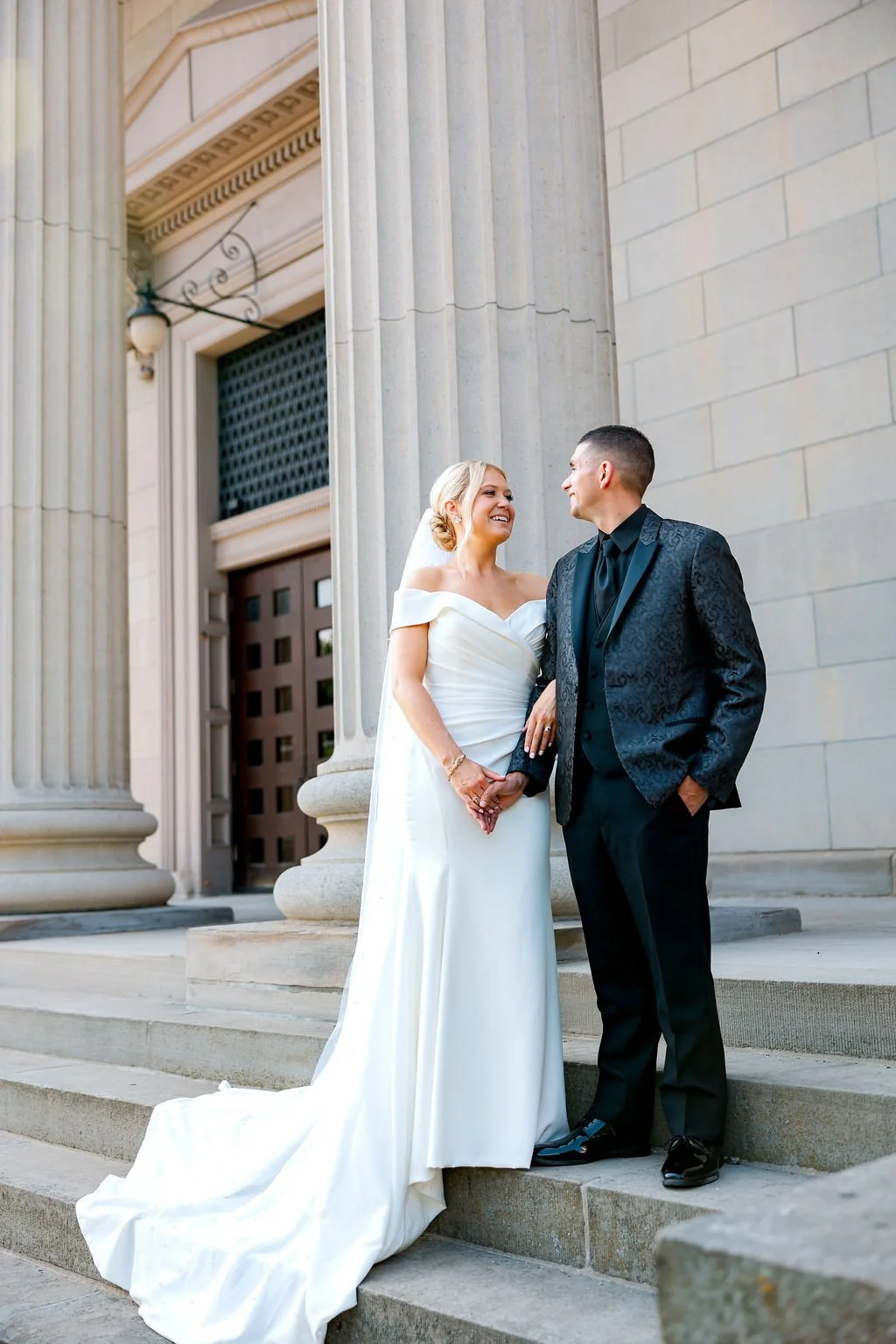 A bride and groom standing on the steps of a government building with large columns, holding hands and smiling at each other.