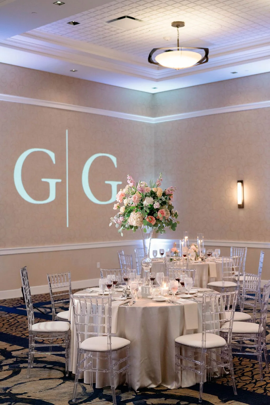 Elegant banquet hall with round table decorated with a tall floral centerpiece, set with glassware, plates, and candles. Clear acrylic chairs surround the table. The wall has a monogram with the letters G and G, and the ceiling features a modern ligh