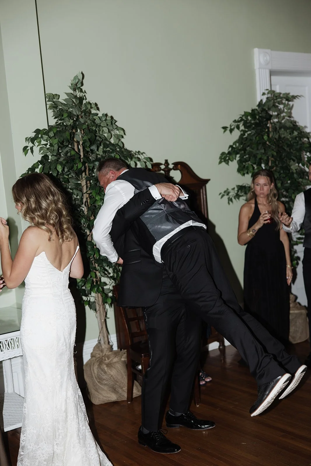 A humorous moment at a wedding reception where a man in a tuxedo is playfully lifting another man in a tuxedo in a hug or dance. The bride in a white wedding dress is standing nearby, and other guests are in the background holding drinks, with plants