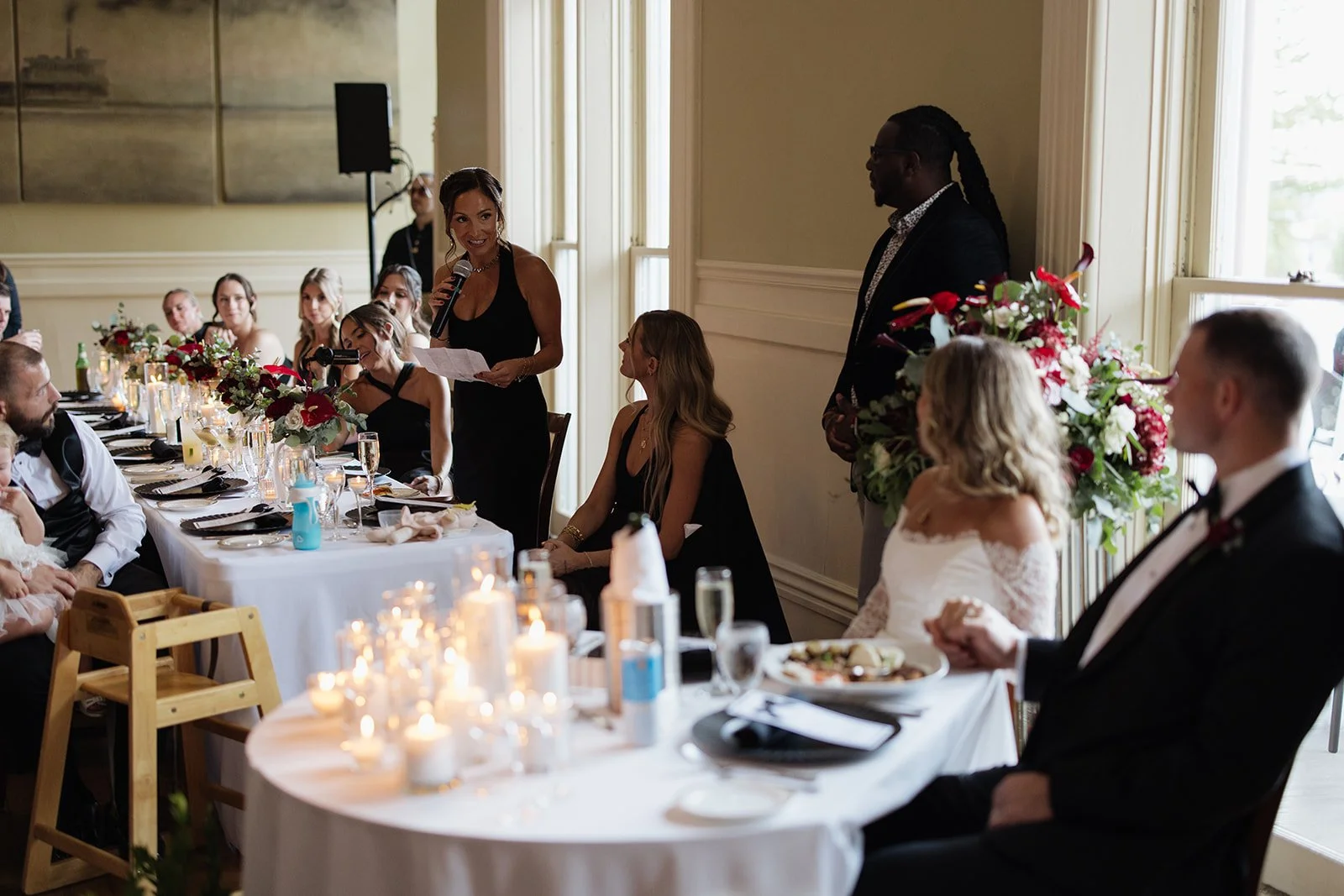A wedding reception with guests seated at a long table decorated with candles and floral centerpieces. A woman is giving a speech with a microphone, while a man stands nearby. The bride and groom are seated at the table, listening to the speech. The 