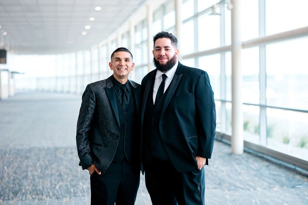 Two men in suits standing in a modern, glass-walled corridor, smiling at the camera.