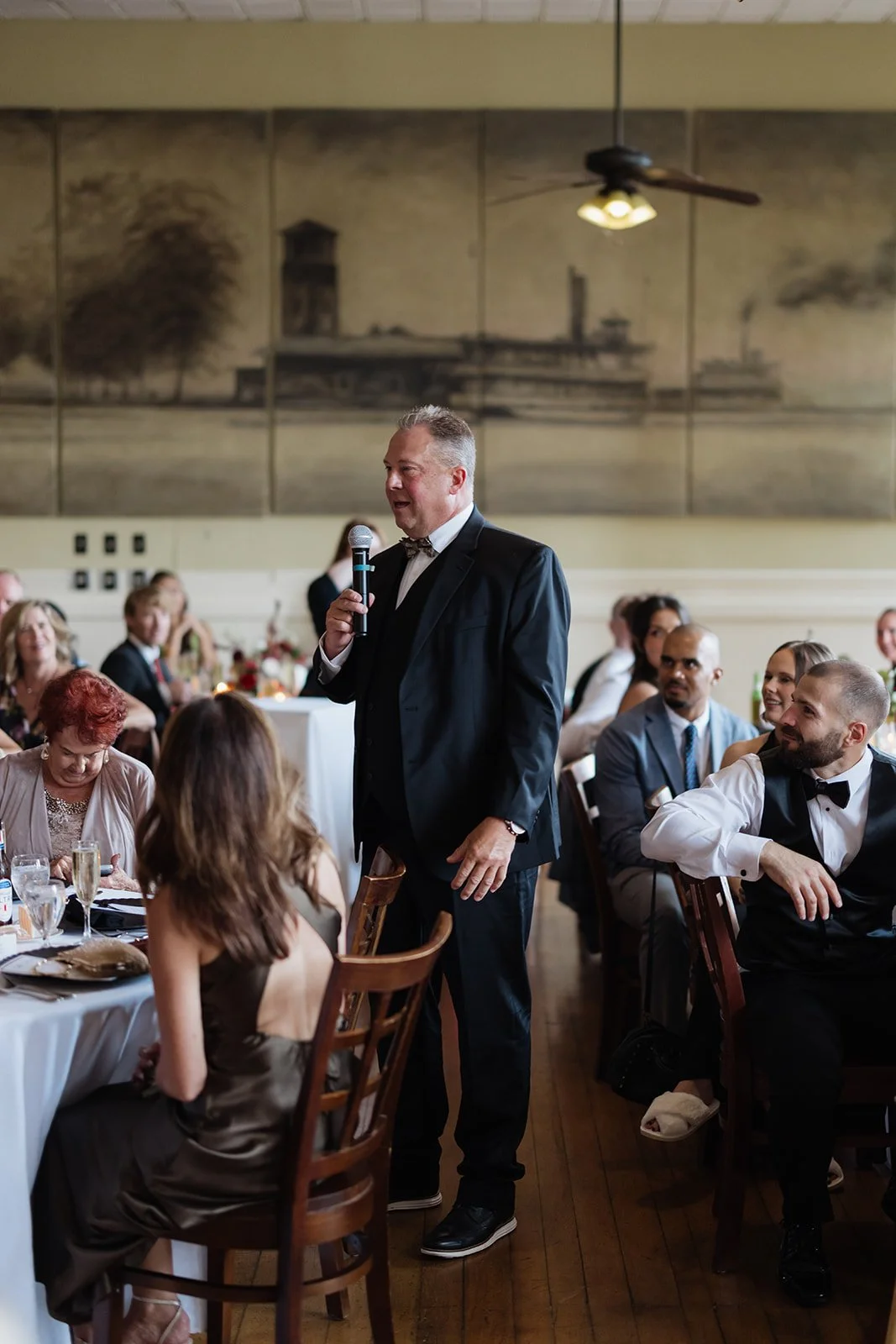 A man in formal attire giving a speech at a wedding reception, surrounded by seated guests.