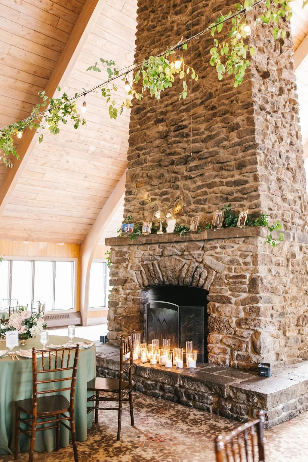 Interior of a rustic lodge or event space with a large stone fireplace, decorated with candles on the hearth, framed photos along the mantle, and string lights with greenery hanging from the ceiling. There is a table set with a green tablecloth and c