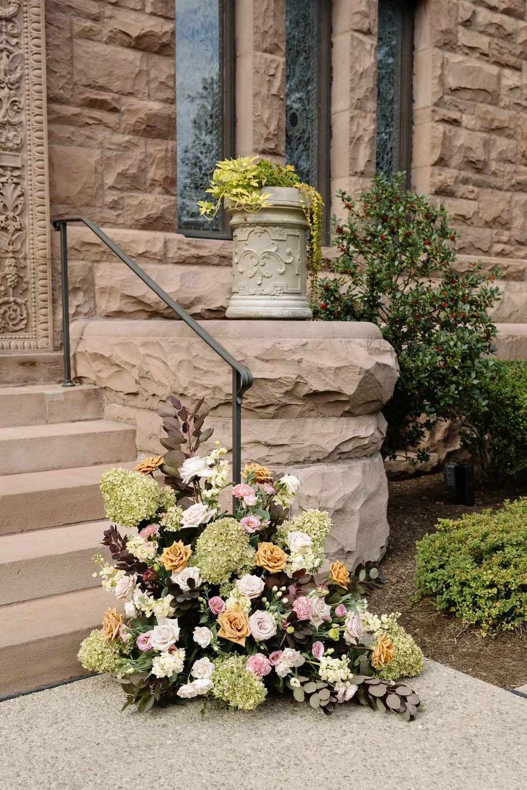 A large floral arrangement with pink, white, and cream-colored flowers is placed at the bottom of a stone staircase outside a building. The building has a brown stone exterior with ornate detailing and a window with decorative glass.