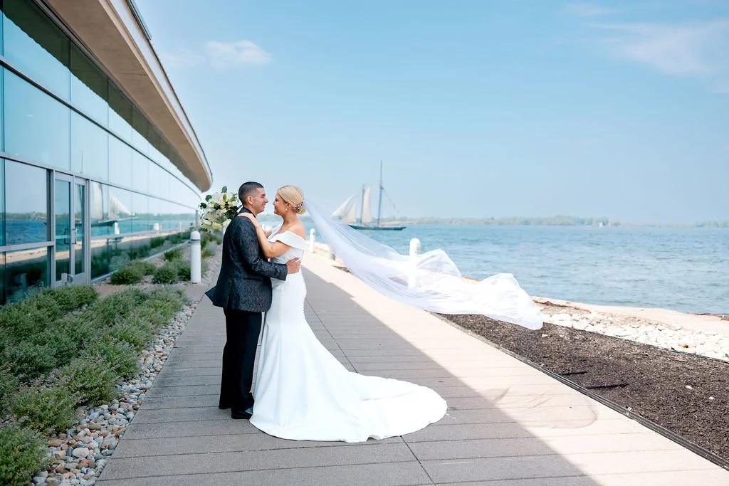 A bride and groom picture by the water, with a sailboat in the background and a long veil flowing in the breeze.
